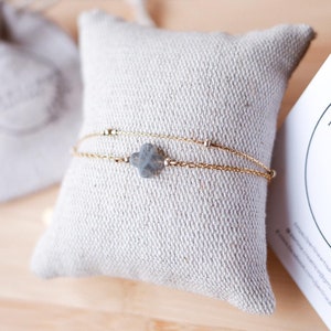 Puede incluir: Una delicada pulsera dorada con un pequeño dije gris de trébol de cuatro hojas descansa sobre una almohada beige. Una pequeña bolsa con cordón y una tarjeta con el texto "PERLINE ARTISANE" están en el fondo. La pulsera es una joya.