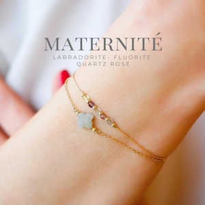 Puede incluir: Una delicada pulsera de cadena dorada de doble hebra con un pequeño dije de flor de piedra de cuatro pétalos y una serie de diminutas cuentas de piedras preciosas multicolores. El texto MATERNITÉ LABRADORITE- FLUORITE QUARTZ ROSE es visible.