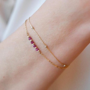Peut inclure: Bracelet doré délicat avec deux fines chaînes. L'une est ornée de petites perles de rubis rondes. L'autre chaîne comporte de petites perles dorées rondes. Le bracelet est porté sur un poignet.