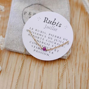 May include: A delicate gold necklace with a small, round, ruby gemstone pendant. The necklace is displayed on a white card with French text, next to a small, beige fabric pouch. The card reads "Rubis Juillet".