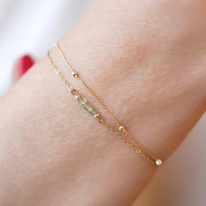 Peut inclure: Un bracelet en or délicat à deux rangs. L'un est orné de petites perles rondes, l'autre de petites perles rectangulaires. Le bracelet est porté au poignet, mettant en valeur ses détails fins et son design élégant.