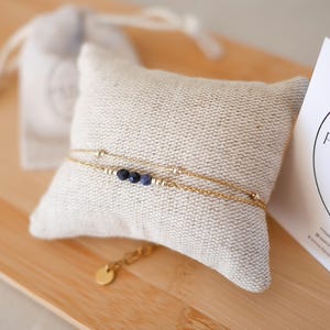 May include: A gold-coloured bracelet with three dark blue beads and small gold beads, displayed on a beige cushion. A small gold charm and a drawstring bag are also visible. The bracelet is presented on a wooden surface.