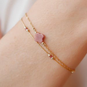 May include: A delicate gold-coloured bracelet with a double chain design. The bracelet features a pink flower-shaped charm and small, red-coloured beads. The jewellery is worn on a wrist, showcasing its elegant and feminine style.