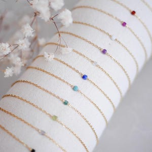 Peut inclure: Une collection de bracelets à chaîne dorée délicats, chacun orné d'une petite perle en pierre précieuse colorée. Les perles varient en couleur, incluant le rouge, rose, blanc, violet, bleu, turquoise, vert et noir, disposées en rangée.
