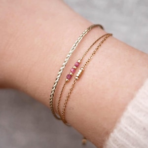 Peut inclure: Trois bracelets dorés portés ensemble. L'un est une chaîne torsadée épaisse. Le deuxième est une chaîne délicate avec trois petites perles roses carrées et des intercalaires dorés. Le troisième est une fine chaîne à maillons avec une petite barre dorée.