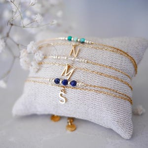May include: Four delicate gold chain bracelets with letter charms and gemstone beads rest on a small, textured pillow. The bracelets feature the letters "N", "M", and "S". The gemstones are turquoise and dark blue.
