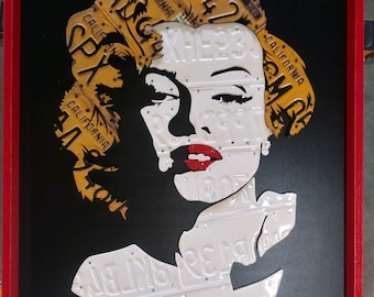 May include: A framed artwork featuring a silhouette of a woman's face created using vintage California license plates. The plates are arranged in a mosaic pattern, with the woman's hair made of yellow plates and her face and body made of white plates. The background is black.