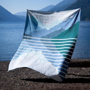 Puede incluir: Un edredón azul y blanco con un diseño de montaña. El edredón presenta un degradado de azules y verdes, con rayas horizontales en la parte inferior. El edredón se exhibe al aire libre cerca de un lago y montañas.