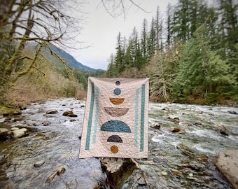 May include: A beige quilt with a geometric design featuring stacked circles and half-circles in shades of brown, teal, and gold. The quilt is displayed outdoors against a backdrop of a river and trees.