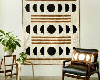 May include: A white quilt with a geometric pattern of black half-circles and brown stripes. The quilt is hanging on a white wall above a black leather armchair with a brown and white patterned pillow.