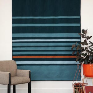 Könnte beinhalten: Eine blaugrüne und orangefarbene gestreifte Steppdecke hängt an einer weißen Wand. Die Steppdecke hat ein geometrisches Muster und wird in einem Raum mit einem braunen Teppich, einem Stuhl und einer Topfpflanze ausgestellt.