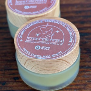 Può includere: Un barattolo di legno con un'etichetta marrone che recita "Divino Femminile, Sensualita, Guarigione, Sacra Dolcezza, Rituale di Autentica Cura di Te, Aromi Mama, Iperico, Canapa, Rosa & Karite".