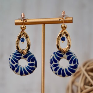 May include: A pair of blue and white ceramic earrings with gold accents hanging from a gold stand. The earrings feature a circular design with a smaller circle in the center.