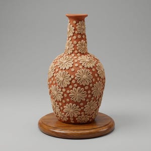 May include: A terracotta-colored vase with a floral pattern. The vase is adorned with raised, cream-colored daisy-like flowers and small dots. It sits on a round, wooden base, showcasing a handcrafted aesthetic. The vase has a long neck and a rounded body.