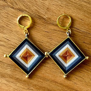 May include: A pair of gold-toned hoop earrings with a diamond-shaped pendant. The pendant features a woven design in black, white, gray, and gold threads. Small gold beads adorn the corners of the diamond.