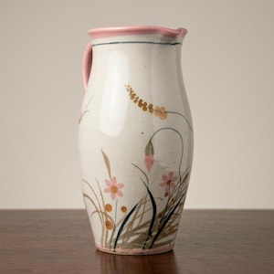 May include: A ceramic pitcher with a pink handle and rim. The off-white pitcher is decorated with pink and gold floral designs and brown and green stems. A thin blue line accents the rim.