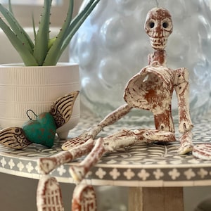 May include: A rustic, hand-painted skeleton figurine with a distressed white and brown finish. The skeleton is seated on a decorative table, with a teal heart-shaped ornament with wings nearby. A white potted plant with green leaves is also visible.