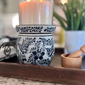 May include: A white ceramic candle holder with a blue and black floral design featuring hummingbirds. The holder has a glass insert for a candle.