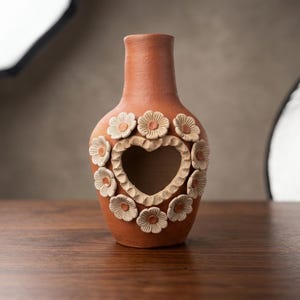 May include: A terracotta-colored clay vase featuring a central heart-shaped cutout surrounded by a decorative border of sculpted, light-colored flowers. The vase has a smooth, matte finish and a narrow neck, with the floral elements arranged in a circular pattern around the heart opening.