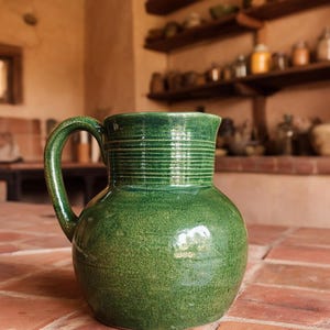 May include: A green ceramic pitcher with a rounded body and a wide, flared rim. The upper neck features a series of horizontal, incised rings, and a single curved handle is attached to the side of the vessel. The glaze has a glossy finish.
