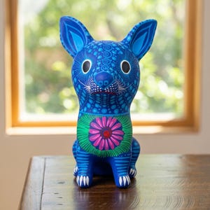 May include: A blue dog figurine featuring intricate patterns of dots and swirls in lighter shades of blue. The chest displays a prominent pink flower with a red center surrounded by green petals. The dog has large ears and white claw details.