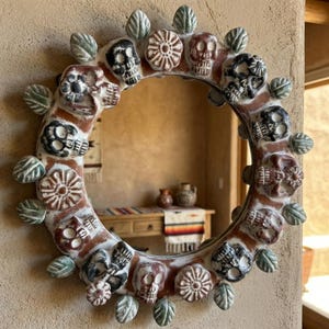 May include: A round decorative mirror with a Day of the Dead theme. The frame is adorned with sculpted skulls, flowers, and leaves in shades of red, white, black, and green. The mirror is mounted on a textured beige wall.