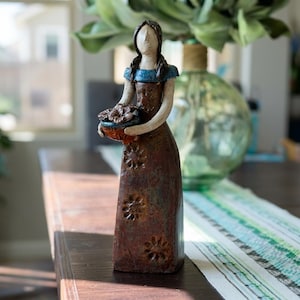 May include: A ceramic figurine of a person with braided hair wearing a blue top and a long brown dress decorated with embossed flower patterns. The figure holds a small bowl containing flowers in both hands, featuring a smooth, faceless head.