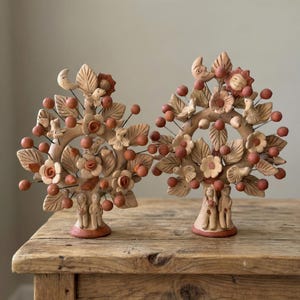 May include: Two ceramic tree sculptures with intricate details. Each tree features flowers, leaves, birds, and red-orange berries. The base of each tree has two figures. The sculptures are displayed on a wooden surface.