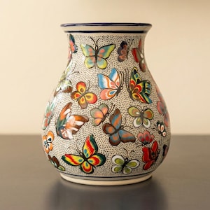 May include: A ceramic vase with a cream-colored background, adorned with colorful butterfly designs. The butterflies feature a variety of colors, including red, yellow, blue, and green. The vase has a dark blue rim and base, and is suitable for home decor.