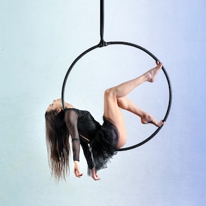 May include: A black aerial hoop suspended by a vertical strap. The hoop is constructed from a circular metal frame wrapped in black tape, providing a grip for aerial performance. The structure is designed to hang from a ceiling point for use.