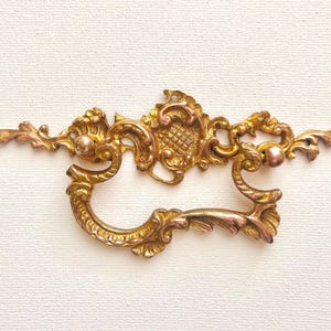May include: Ornate gold-toned metal drawer pull with a decorative, intricate design. The pull has a ring-shaped opening and two decorative knobs.