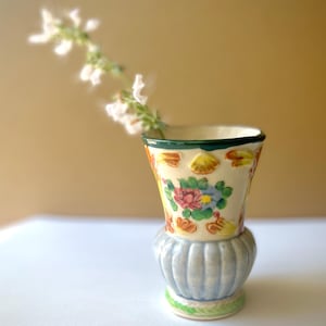 May include: A small ceramic vase with a flared rim and a ribbed, light blue base. The upper section is cream-colored with hand-painted floral motifs in pink, green, and orange. A single sprig of small white flowers extends from the vase opening.