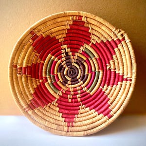 May include: A round, shallow woven basket featuring a coiled design. The basket is constructed from natural tan fibers accented with concentric patterns in vibrant red, dark brown, and muted green, creating a geometric, radial visual effect.