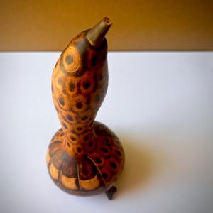 May include: A gourd sculpture shaped like a bird with a long neck and a pointed beak. The surface features a natural tan and brown color palette with dark circular patterns and segmented markings covering the rounded body and the curved neck.