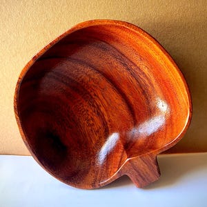 May include: A polished wooden bowl with a smooth, rounded shape and a distinct, short handle extending from one side. The bowl features a rich, warm reddish-brown color with visible natural wood grain patterns throughout the interior surface.