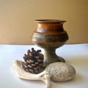 May include: A ceramic chalice with a flared rim, featuring a glossy orange upper section and a mottled brown and green base. Beside it sits a brown pinecone, a smooth grey speckled river stone, and a curved, off-white animal bone fragment.