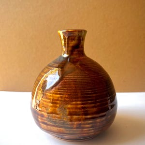 Könnte beinhalten: Eine kleine, runde Keramikvase mit engem Hals und einer glänzenden, dunkelbraunen Glasur. Die Oberfläche weist horizontale Rillen auf, die den Körper umkreisen, mit helleren bernsteinfarbenen Tropfen und Streifen, die vertikal verlaufen.