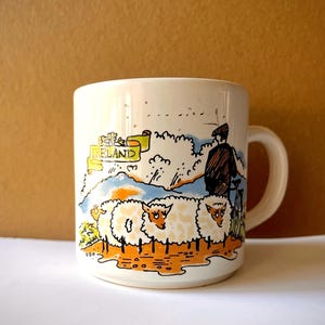 May include: A white ceramic mug featuring a colorful illustration of two fluffy sheep standing in a field with a person on a bicycle in the background. A yellow banner with the text IRELAND is printed above the sheep, set against a landscape.