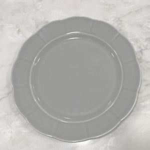 May include: A pale grey ceramic plate with a scalloped edge. The plate has a smooth, glossy finish and a slightly raised rim. The plate is round and appears to be new.