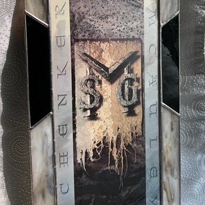 May include: A stained glass framed picture with a black and white image of a band logo. The logo is a stylized "SG" with a dollar sign and a sword. The frame is made of black and white glass with a beige accent.