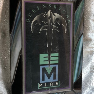 Puede incluir: Una imagen enmarcada en negro con un borde de vidrio tintado verde y azul. La imagen presenta un gráfico en blanco y negro de una espada estilizada con el texto "Queensrÿche" y "Empire" en blanco.