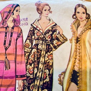 May include: A vintage sewing pattern envelope featuring illustrations of three different robe styles. The left robe is pink and orange with a hood and tassel. The center robe has a dark brown floral pattern. The right robe is yellow with pom-pom trim.