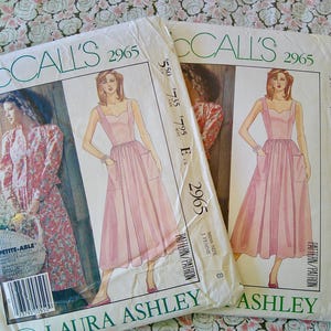 May include: Two vintage McCall's sewing patterns, featuring illustrations of dresses. The patterns are labeled "2965" and include the text "Laura Ashley." The dresses are light pink with pockets and straps. The background is a floral pattern.