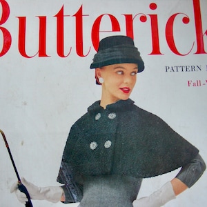 May include: Vintage Butterick Pattern Book cover featuring a woman in a dark cape and hat, holding a curved handle umbrella. The cover text reads "Butterick" in red, with "Pattern Book Fall-Winter 1956".