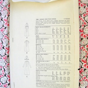 Op de afbeelding: Een vintage naaipatroon voor een eendelige jurk met een afneembare kap. Het patroon is voor maten 6-14 en bevat instructies voor het maken van de jurk met of zonder pool. Het patroon is gedrukt op wit papier met zwarte tekst en illustraties.