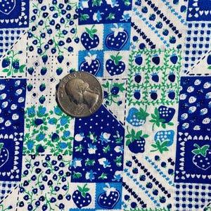 Op de afbeelding: Close-up van een stof met een patchwork-ontwerp met aardbeien en bosbessen in blauwe en groene tinten op een witte achtergrond. De stof heeft een vintage, cottagecore esthetiek.