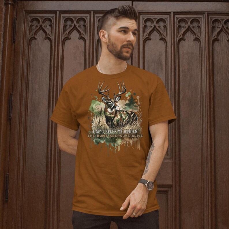 May include: A brown t-shirt featuring a camouflage print of a deer and the text "CAMO KEEPS ME HIDDEN THE HUNT KEEPS ME ALIVE".