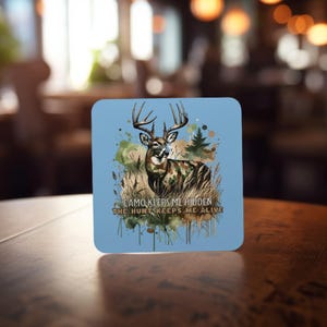 May include: A blue square coaster featuring a watercolour illustration of a white-tailed deer in camouflage standing in a woodland setting. The text "CAMO KEEPS ME HIDDEN THE HUNT KEEPS ME ALIVE" is printed below the deer.