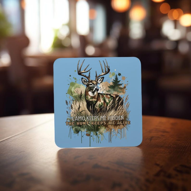 May include: A blue square coaster featuring a watercolour illustration of a white-tailed deer in camouflage standing in a woodland setting. The text "CAMO KEEPS ME HIDDEN THE HUNT KEEPS ME ALIVE" is printed below the deer.