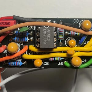 以下が含まれることがあります： 様々な電子部品が実装された黒色の回路基板のクローズアップ。基板には「LM4250CJ 7910」とラベル付けされた集積回路、抵抗器、コンデンサ、および色付きのワイヤが含まれています。「473J100」の文字も確認できます。